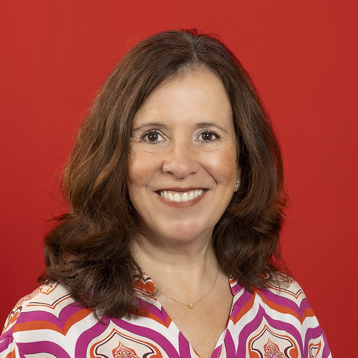 Foto-zeigt-Porträt-einer-Frau-mit-schulterlangem-braunem-Haar-vor-rotem-Hintergrund-mit-freundlichem-offenem-Blick-in-die-Kamera