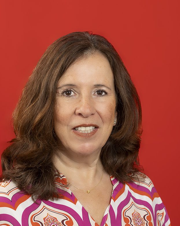 Foto-zeigt-Porträt einer Frau mit schulterlangem braunem Haar vor rotem Hintergrund, freundlich lächelnd, in gemusterter Bluse.